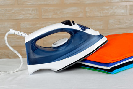 New modern electric steam iron and a stack of ironed t-shirts of various colors on ironing board on...