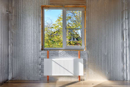 Metal heating radiator in a country house under construction. White radiator under the window against the background of walls sheathed with heat-insulating materialの写真素材
