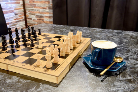 Chess board with figures and blue cup with cappuccino coffee on marble table against brown curtain...