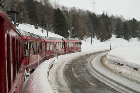 SUROVAS, SWITZERLAND - January, 2018. The train Bernina Express,  in a passage near the road, crosses the Swiss alps in winter with snowのeditorial素材