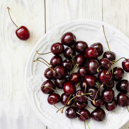 Dark sweet cherry in a white plate and on light wooden table close-up. Top view.