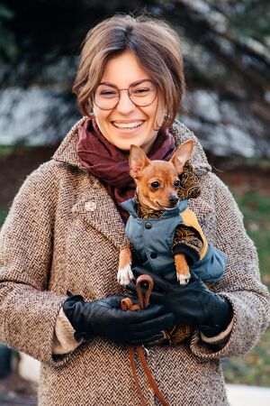 smiling girl in coat holds a small dog toy Terrier in her arms in autumn.の写真素材
