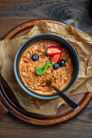 Homemade creme brulee with strawberries and mint in a metal bowl with a spoon on parchment on a dark...