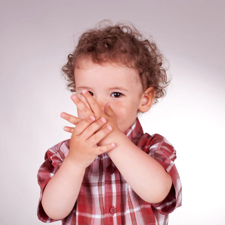 Little curly boy holding his hands in front of the face and facing the camera, on a gray background