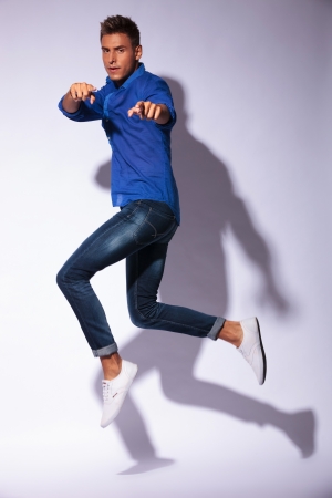 full length picture of a young casual man jumping in the air and pointing at the camera with both hands, on gray background with shadowの写真素材