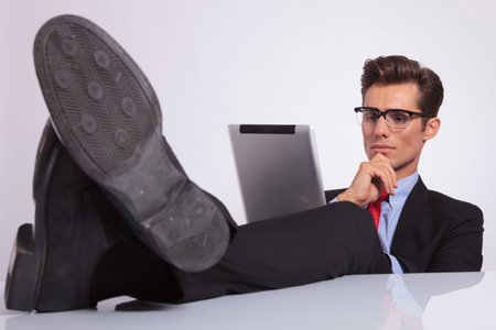 young business man sitting with legs crossed on the desk and looking thoughtfully at his tablet, on...