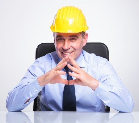 senior bussines man with helmet sitting at the desk with his hands together and smiling at the camera. on gray background の写真素材