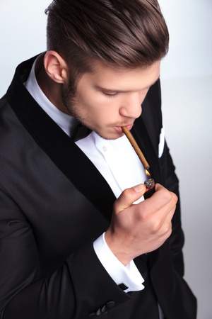 closeup picture of an elegant young fashion man in tuxedo lighting up his cigar. on gray backgroundの写真素材