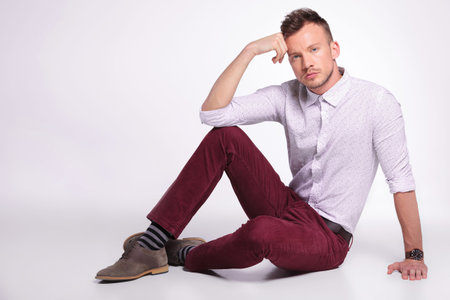 casual young man posing on the floor with his hand at his temple and elbow on knee while looking at...