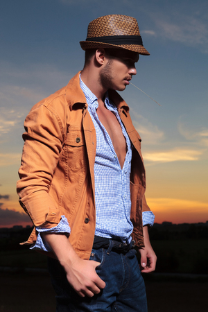 side view of a casual young man standing outdoor with a straw in his mouth and a thumb in his pocket while looking away from the camera with the sunset behind himの写真素材
