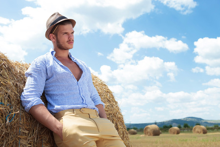 young casual man posing outdoor with both hands in pockets and looking away while leaning back on a haystackの写真素材