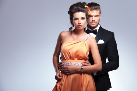 young fashion couple with man behind woman holding her with both hands while both looking at the camera. on gray backgroundの写真素材