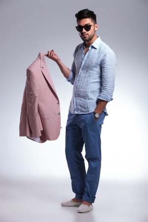 full length picture of a young casual man holding his jacket on his finger and a hand in his pocket while looking into the camera. on gray backgroundの写真素材