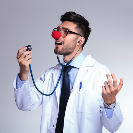 young male doctor listening himself sing at the stethoscope while wearing a clown red nose. on gray backgroundの写真素材