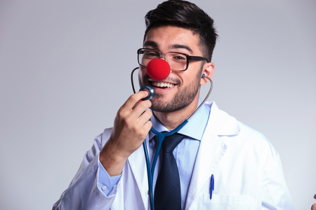 young male doctor with a red clowns nose listening to himself at the stethoscope and smiling for the camera. on gray backgroundの写真素材