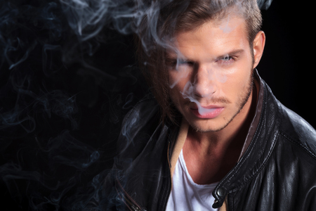 young fashion man in leather jacket covered in smoke on black backgroundの写真素材