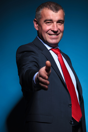 old smiling business man welcomes you with a handshake on blue backgroundの写真素材