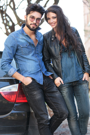 young casual couple standing by their car, with their hands in the pockets while smiling for the cameraの写真素材