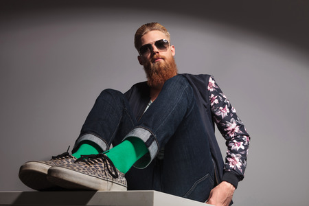 angle view of a casual young man with a long red beard sitting on a table and looking down, into the camera. in a gray background studioの写真素材