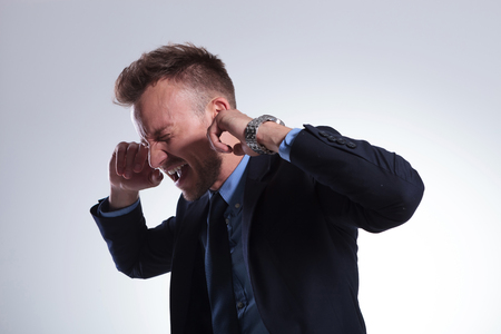 young business man plugging his ears with his fingers and closing his eyes while screaming. on a light gray studio backgroundの写真素材