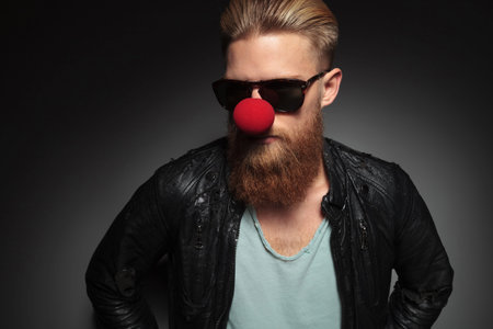casual young man with a long beard, wearing a clown's red nose and looking away . on a dark studio...