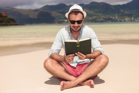 smiling young man reading a good book while sitting on the beachの写真素材