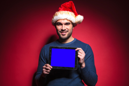 Young santa man holding a tablet pad computer with a blue screen, smiling for the camera.の写真素材
