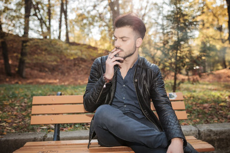 Young fashion man sitting on a bench in the park enjoying his cigarette.の写真素材