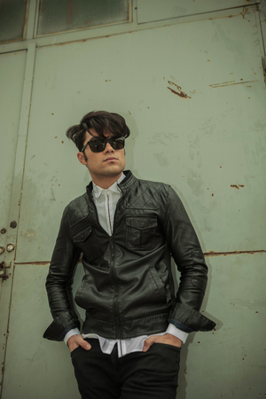 Down view of a young fashion man holding both hands in pocket while leaning on a green metal door.の写真素材