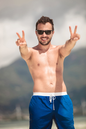 Portrait of a young happy man posing on the beach while showing the victory sign with both hands.の写真素材