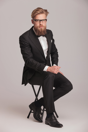 Handsome young business man looking at the camera while sitting on a stool, holding one hand on his knee.の写真素材