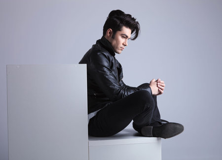 Side view of a fashion man sitting on a while furniture while looking away from the camera with his hands together.の写真素材