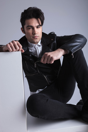 Handsome fashion man sitting on the floor while holding one hand on a white table.の写真素材