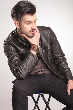 Close up picture of a young fashion man resting on a stool while looking away and holding his hand to the chin.の写真素材