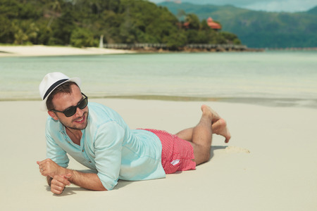 smiling man lying in the sand with his hands crossed while looking away from the cameraの写真素材