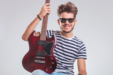 young rocker posing in studio background with electric guitar in his lap. he is seated, wearing sunglasses and is looking at the camera.の写真素材