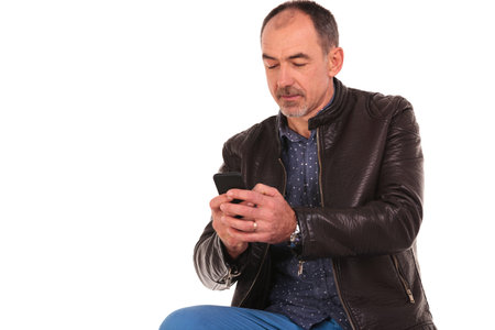 casual bearded man in leather jacket posing seated in isolated studio background while texting on...