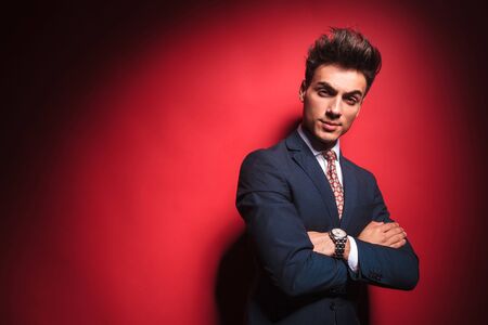 portrait of confident young businessman in black suit with red tie posing with hands crossed while frowning and looking at the camera in red studio backgroundの写真素材