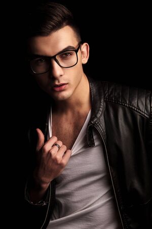 closeup portrait of sexy man in black leather jacket and glasses, pulling his white shirt while looking at the camera in isolated black studio backgroundの写真素材