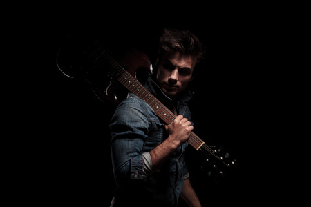 dramatic young guitarist looking back while holding guitar on shoulder, on black studio backgroundの写真素材