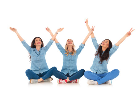 three happy women making the victory hand sign while sitting and look up at somethingの写真素材