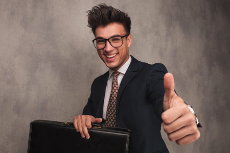 happy young business man sitting with a briefcase and makes the ok thumbs up hand sign in studio