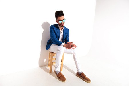 side of a fashion man sitting on chair and rubbing his hands while looking away from the camera on white backgroundの写真素材