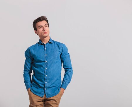 casual young man with hands in pockets looks away to side, thinking on grey backgroundの写真素材