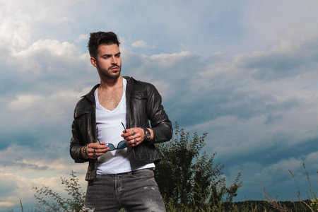 handsome man in leather jacket holding sunglasses looks to side in the outdoors