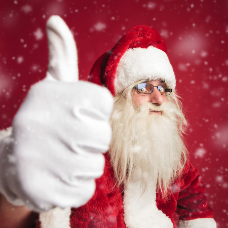 excited santa claus making the ok thumbs up hand sign on red background with snow fallingの写真素材