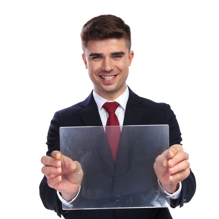 portrait of excited businessman presenting blank transparent futuristic gadget while standing on white background. He wears a navy coloured suit and a red tie.の写真素材