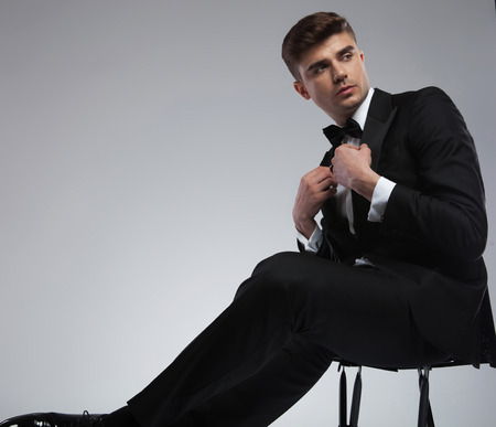 portrait of handsome elegant young man fixing suit collar and looking to side while sitting on a metal chair on light grey backgroundの写真素材