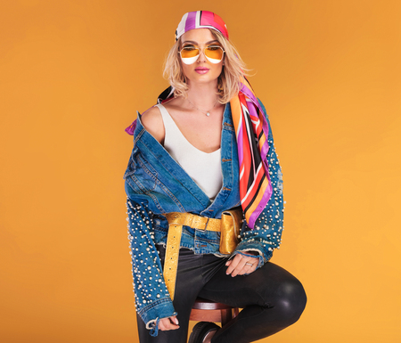 beautiful blonde woman wearing colored clothes sitting on wooden chair on yellow backgroundの写真素材