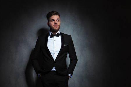 elegant young man standing with hands in pockets looks up to side near a dark grey wallpaper. He is wearing a black tuxedo, looking pensiveの写真素材